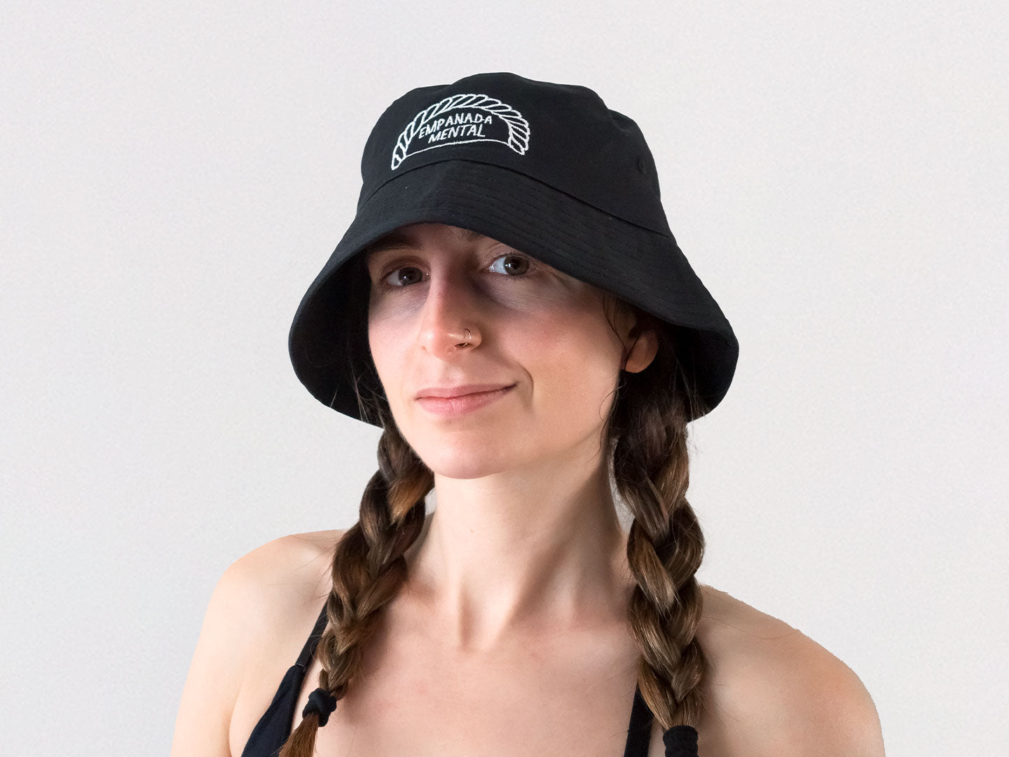 Chica con trenzas que lleva un gorro de pescador bucket hat negro con un bordado de una ilustración de una empanadilla en la que pone 