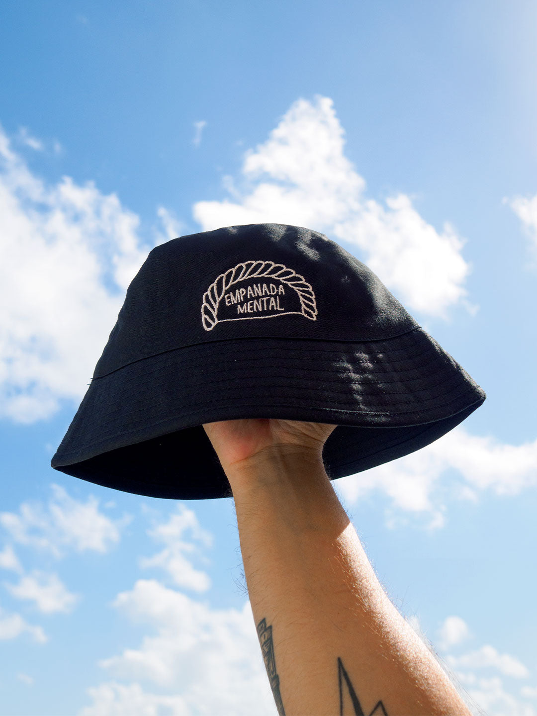 Mano agarrando un gorro de pescador bucket hat negro con bordado de una ilustración de una empanadilla en la que pone 