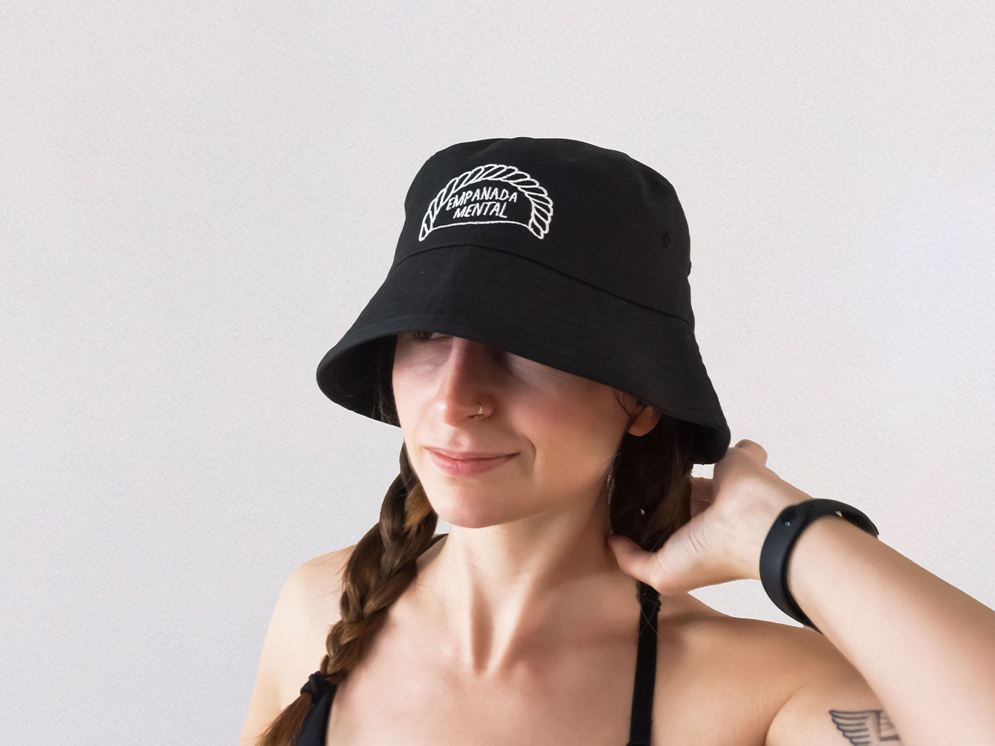 Chica con trenzas que lleva un gorro de pescador bucket hat negro con un bordado de una ilustración de una empanadilla en la que pone 