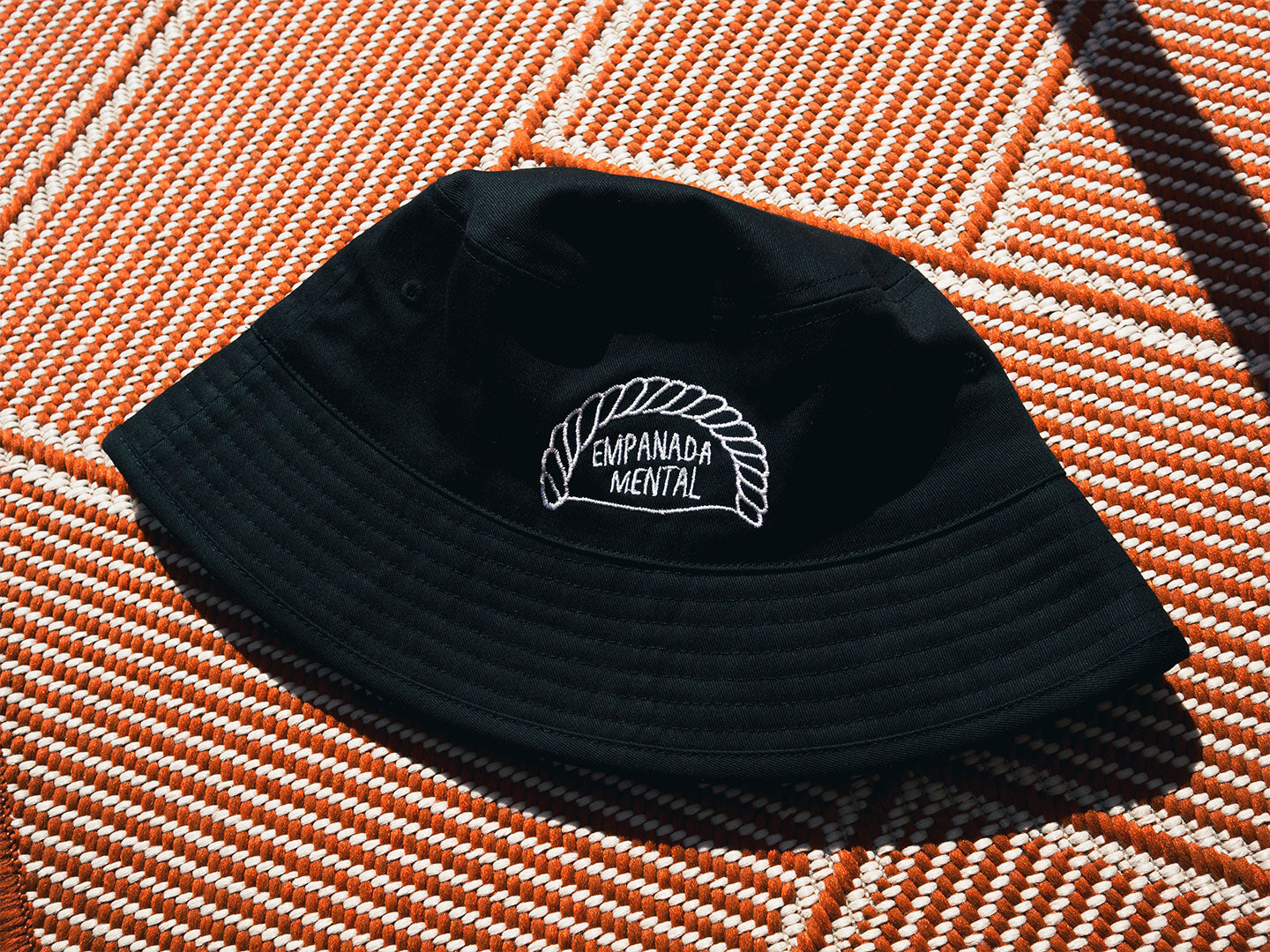 Gorro Bucket hat negro sobre un fondo color terracota. En el gorro hay una ilustración bordada de una empandilla en la que pone 